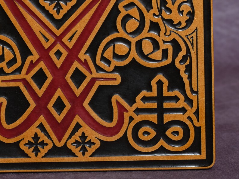 May include: A black leather square with a red and brown design. The design features a cross with a circle around it, and a symbol with two intersecting lines.