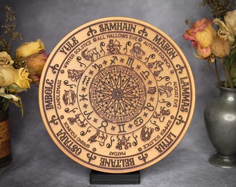 May include: A round, wooden decorative piece with engraved zodiac symbols and seasonal references. The light brown surface features intricate details, including astrological signs and text such as "Yule" and "Samhain". The piece is displayed on a black stand.