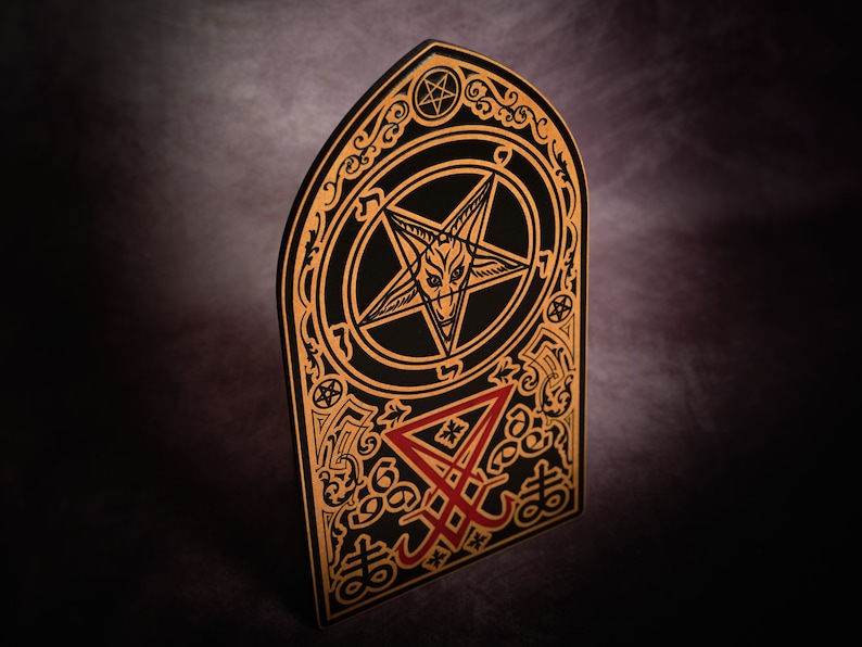 May include: A black and gold wooden plaque featuring a pentagram with a goat's head inside. The plaque also has a red inverted pentagram with the word "666" at the bottom.