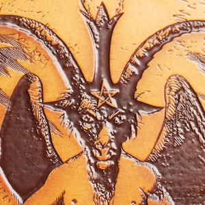 May include: A brown leather embossed image of a goat with horns and wings. The goat has a pentagram on its forehead and a beard. The background is a light brown color.