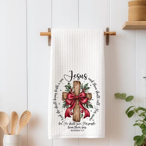 May include: White waffle-weave kitchen towel with a printed design featuring a wooden cross adorned with a red bow and holly. The word "Jesus" is at the top, with a Bible verse encircling the cross.