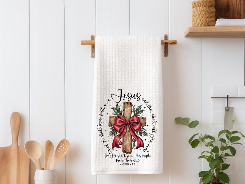 May include: White waffle-weave kitchen towel with a printed design featuring a wooden cross adorned with a red bow and holly. The word "Jesus" is at the top, with a Bible verse encircling the cross.