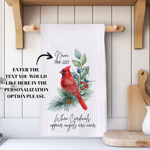 May include: White waffle-weave kitchen towel with a watercolor cardinal design. The towel features the text "Nana 1948-2023" and "When Cardinals appear angels are near." The design includes a red cardinal, greenery, and berries.