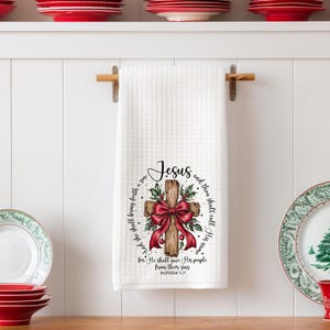 May include: White waffle-weave kitchen towel with a wooden cross design. The cross is adorned with a red bow and holly. The towel has the word "Jesus" and a Bible verse printed on it. Red and green plates are on the shelf.
