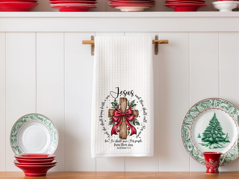 May include: White waffle-weave kitchen towel with a wooden cross design. The cross is adorned with a red bow and holly. The towel has the word "Jesus" and a Bible verse printed on it. Red and green plates are on the shelf.