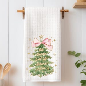 May include: White waffle-weave kitchen towel with a Christmas tree design. The tree is green with a pink bow and gold accents. Text on the towel reads "Oh Come Let Us Adore Him". The towel is hanging on a wooden rod.