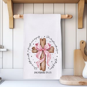 May include: White waffle-weave kitchen towel with a wooden cross design. The cross is adorned with a pink bow and surrounded by text. The towel is hanging on a wooden rod in a kitchen setting.