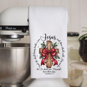 May include: White kitchen towel with a printed design featuring a wooden cross, a red bow, holly, and the word "Jesus." The towel has text from Matthew 1:21. The towel is hanging on a white stand mixer.