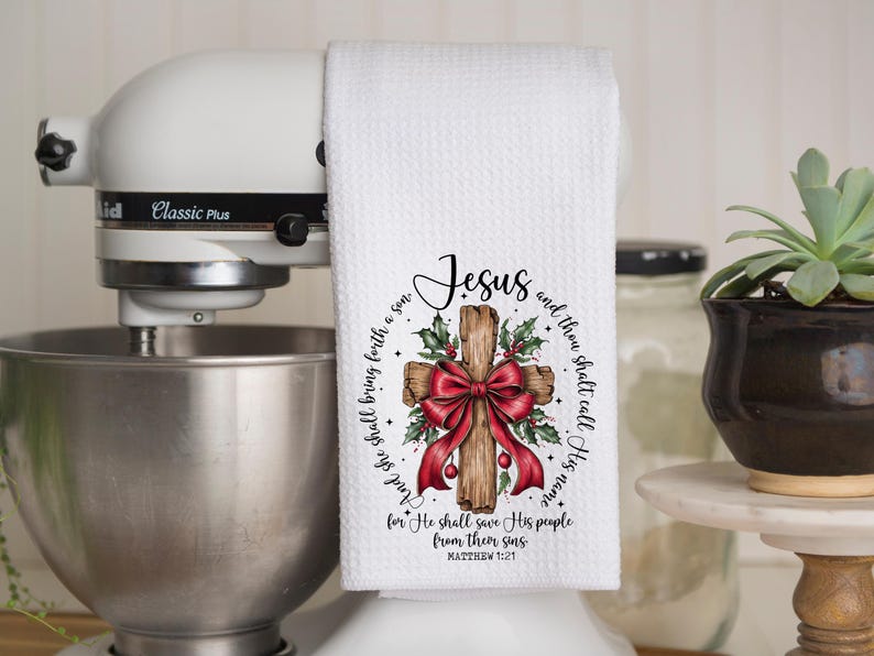 May include: White kitchen towel with a printed design featuring a wooden cross, a red bow, holly, and the word "Jesus." The towel has text from Matthew 1:21. The towel is hanging on a white stand mixer.