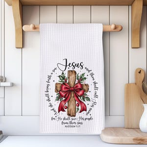 May include: White waffle-weave kitchen towel with a printed design featuring a wooden cross adorned with a red bow and holly. The text "Jesus" is above the cross, with a Bible verse encircling it. A decorative kitchen accessory.