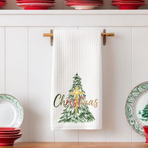 May include: White waffle-weave kitchen towel with a Christmas tree and cross design. The towel has the word "Christmas" in green and gold script. Red and green plates and a red cup are in the background.