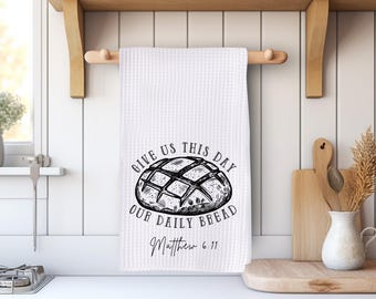 May include: White waffle-weave kitchen towel with a black illustration of a loaf of bread and the text "GIVE US THIS DAY OUR DAILY BREAD Matthew 6:11". The towel hangs on a wooden rod.