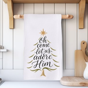 May include: White waffle-weave kitchen towel with a Christmas tree design and the text "Oh come let us adore Him" in black script. The tree is outlined in green with a gold star and base. The towel hangs on a wooden rod.