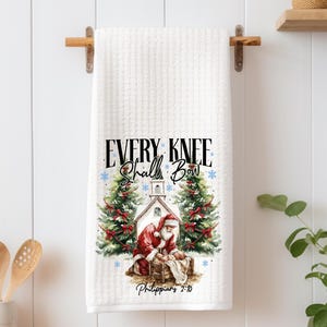 May include: White waffle-weave kitchen towel with a Christmas-themed graphic. The graphic features Santa Claus, a church, and Christmas trees. Text on the towel reads "EVERY KNEE Shall Bow" and "Philippians 2:10". The towel is hanging on a wooden rod.