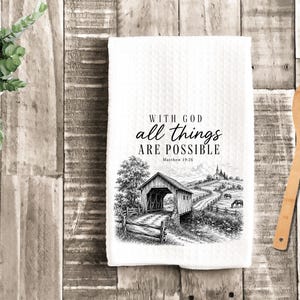 May include: A white waffle-weave kitchen towel featuring a black ink illustration of a covered bridge, a winding road, and a church. The text reads: WITH GOD all things ARE POSSIBLE Matthew 19:26. The design includes trees and horses.