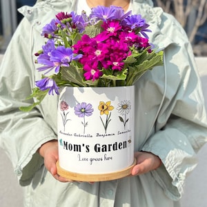 May include: A white ceramic flower pot with a floral design and the text "Mom's Garden". The pot holds a vibrant bouquet of purple and pink flowers. The pot sits on a round wooden base. The text "Love grows here" is also on the pot.