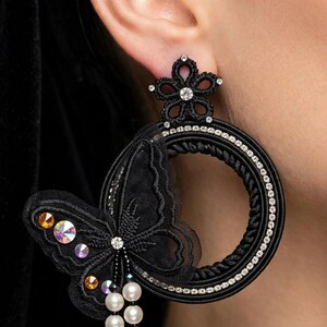 Può includere: Un orecchino a cerchio nero caratterizzato da un perno a forma di fiore con un cristallo centrale, un grande telaio circolare decorato con strass e un dettaglio a farfalla in pizzo nero impreziosito da cristalli iridescenti e tre perle bianche.