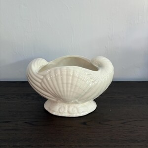 May include: A glossy off-white ceramic bowl shaped like a large seashell. The vessel features intricate, raised radial ridges mimicking the texture of a scallop shell, with two prominent curled edges at the top and a sturdy, circular pedestal base.