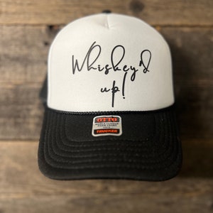 May include: White trucker hat with black mesh back and a black bill. The hat has the text "Whiskey'd up!" written in black cursive on the front. The hat has a black and orange "OTTO" label on the side.