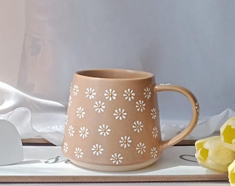 Op de afbeelding: Een bruin keramische mok versierd met witte madeliefjespatronen. De mok heeft een gebogen handvat en staat op een wit dienblad. Gele tulpen en een witte hartvormige decoratie zijn ook aanwezig.