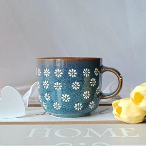 May include: A blue ceramic mug with a daisy pattern. The mug has a brown rim and handle, and is decorated with white daisies with yellow centers. The mug is sitting on a white tray with the word "HOME" stenciled on it.