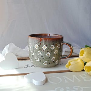 May include: A ceramic mug with a green and brown glaze, adorned with white daisy cutouts. The mug has a brown handle and rim. The mug is on a white surface with heart-shaped decorations and yellow tulips.