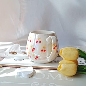 May include: A pearlescent white ceramic mug adorned with red cherry illustrations. The mug has a curved handle and a rounded body. Yellow tulips and decorative elements are in the background.