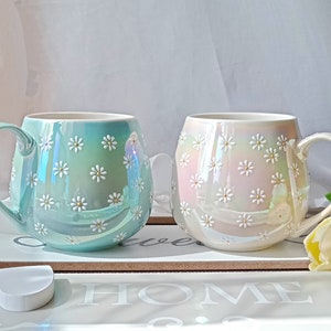 May include: Two ceramic mugs with a daisy pattern. The mug on the left is light blue with a pearlescent finish. The mug on the right is white with a pearlescent finish.
