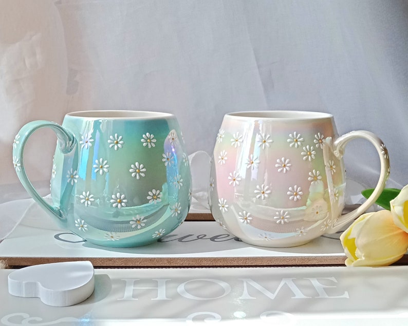 May include: Two ceramic mugs with a daisy pattern. The mug on the left is light blue with a pearlescent finish. The mug on the right is white with a pearlescent finish.