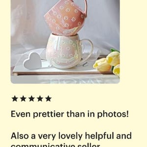 May include: Two pink ceramic mugs with a daisy pattern. The mugs are stacked on top of each other. The mug on top is slightly tilted. The mugs are on a white surface with a white heart and yellow flowers.