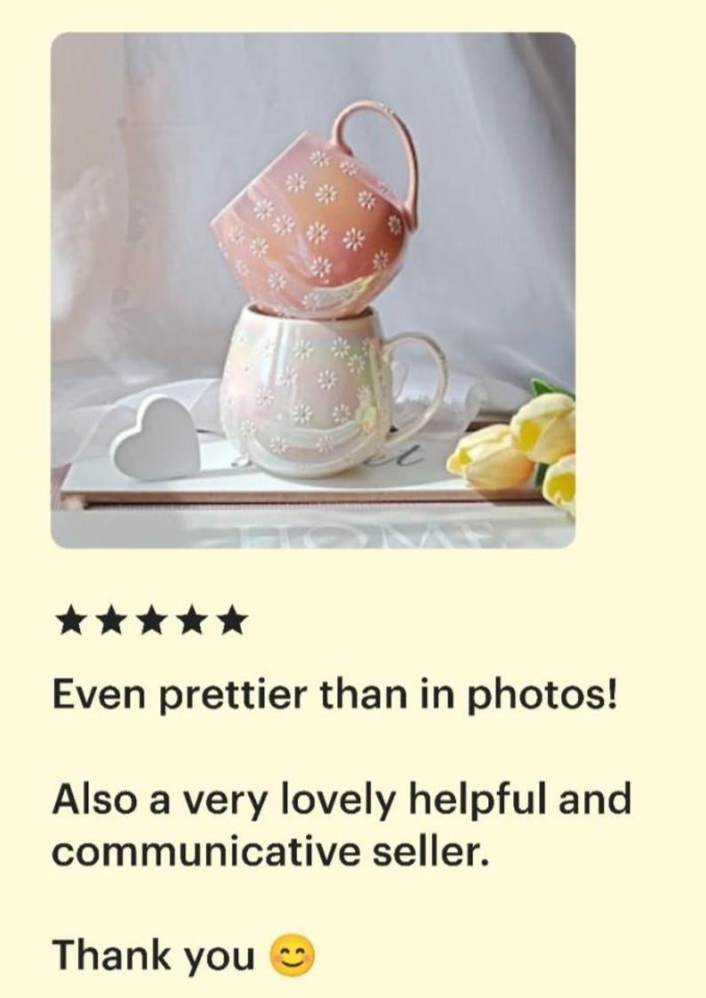 May include: Two pink ceramic mugs with a daisy pattern. The mugs are stacked on top of each other. The mug on top is slightly tilted. The mugs are on a white surface with a white heart and yellow flowers.