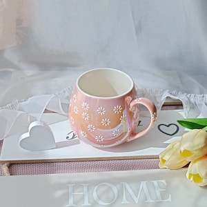 May include: A pink ceramic mug with a white daisy pattern. The mug has a handle and is sitting on a wooden surface with a white heart and the word "HOME" written on it.