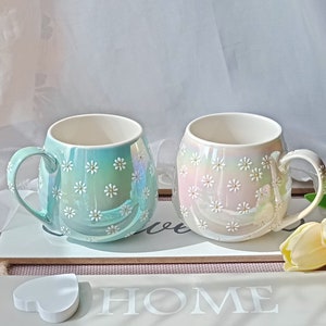 May include: Two ceramic mugs with a daisy pattern. The mug on the left is light blue with white daisies. The mug on the right is white with pink and white daisies. The mugs are sitting on a wooden surface with the word "HOME" written on it.
