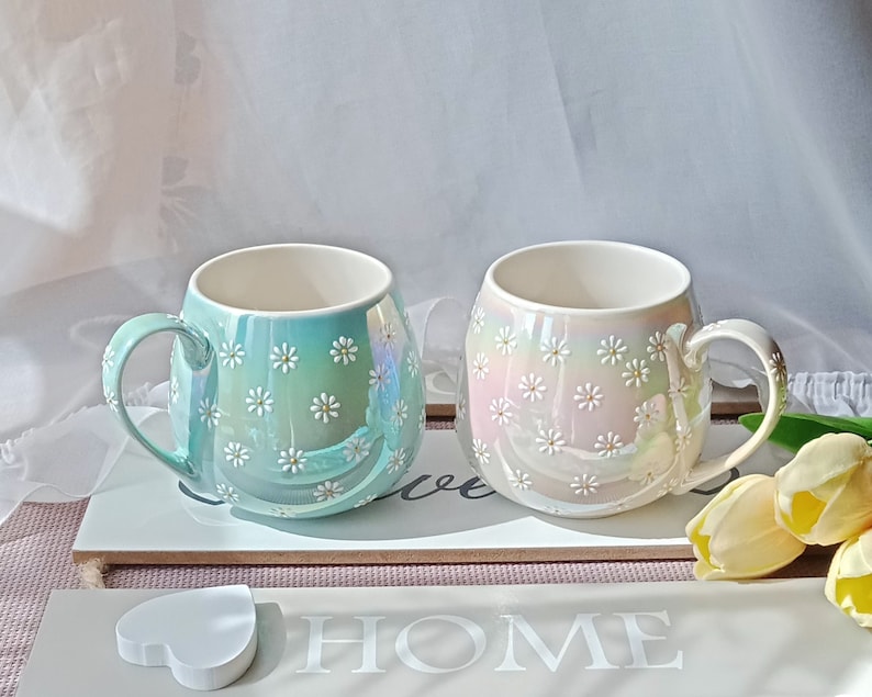May include: Two ceramic mugs with a daisy pattern. The mug on the left is light blue with white daisies. The mug on the right is white with pink and white daisies. The mugs are sitting on a wooden surface with the word "HOME" written on it.
