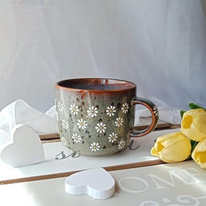 May include: A ceramic mug with a floral daisy pattern. The mug has a dark green base with white daisies and a brown rim and handle. Two white wooden heart decorations and yellow tulips are also in the image.