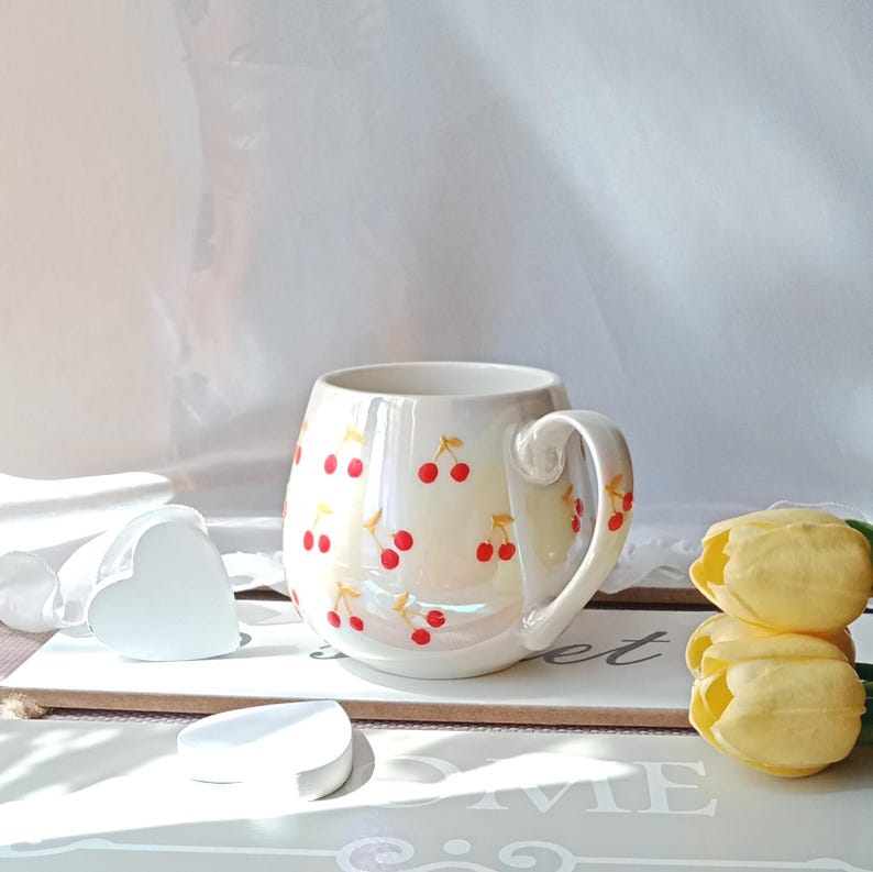May include: A white ceramic mug with a rounded shape and a large handle. The mug is decorated with red cherry illustrations. The mug is sitting on a white surface with a heart-shaped object and yellow tulips.