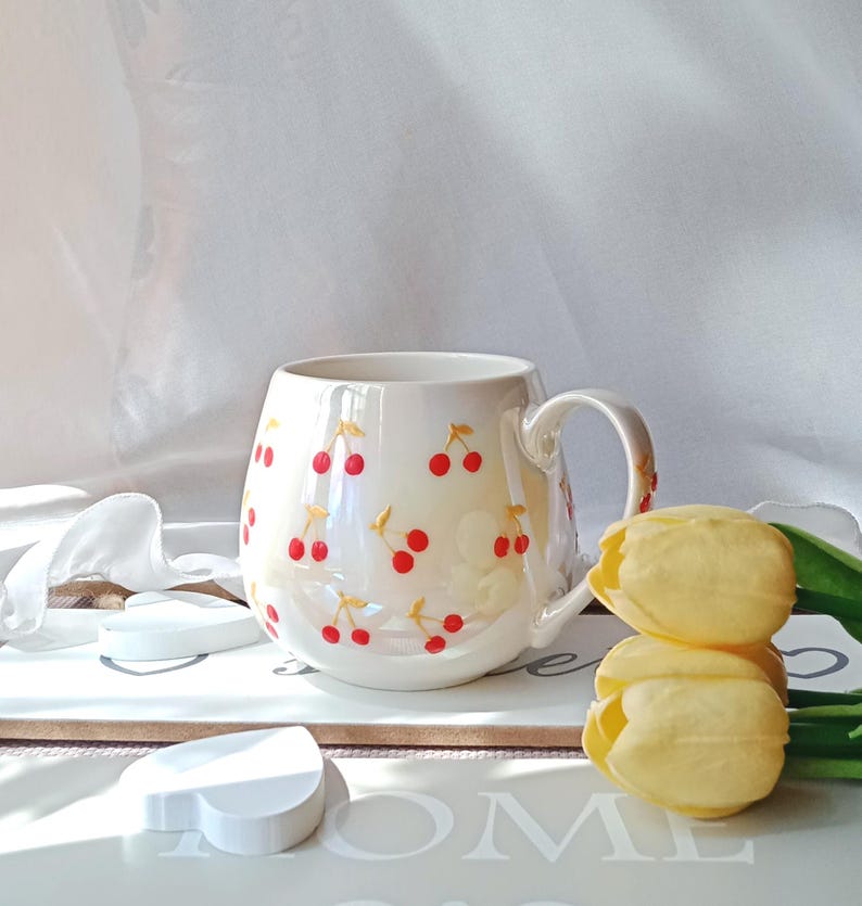 May include: A white ceramic mug with a rounded shape and a handle. The mug is decorated with red cherries and gold stems. The mug is sitting on a white tray with a heart-shaped object and yellow tulips.