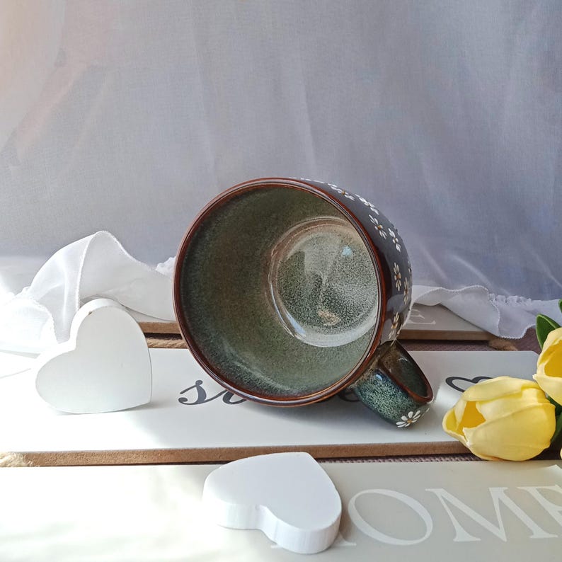 May include: A dark brown ceramic mug with a green interior and a floral pattern. The mug is decorated with small white flowers and has a matching handle. Two white heart-shaped decorations are in the foreground.
