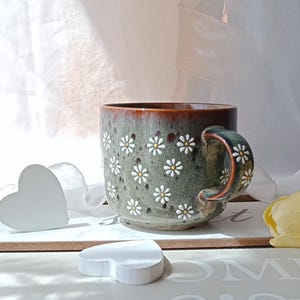 May include: A ceramic mug with a floral daisy pattern. The mug has a green and brown glaze with white and yellow daisy accents. The handle is also decorated with daisies. The mug is sitting on a white surface.