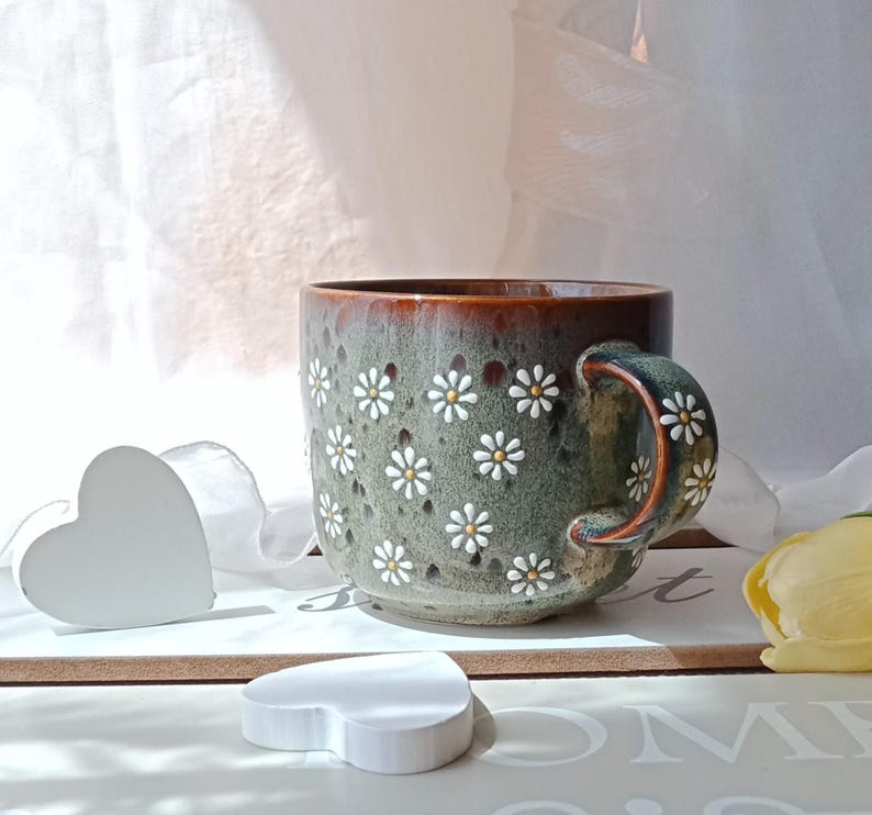May include: A ceramic mug with a floral daisy pattern. The mug has a green and brown glaze with white and yellow daisy accents. The handle is also decorated with daisies. The mug is sitting on a white surface.