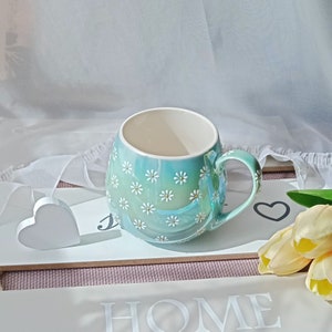 May include: A light blue ceramic mug with a white daisy pattern. The mug has a rounded shape and a handle. The mug is sitting on a white surface with a heart-shaped decoration.