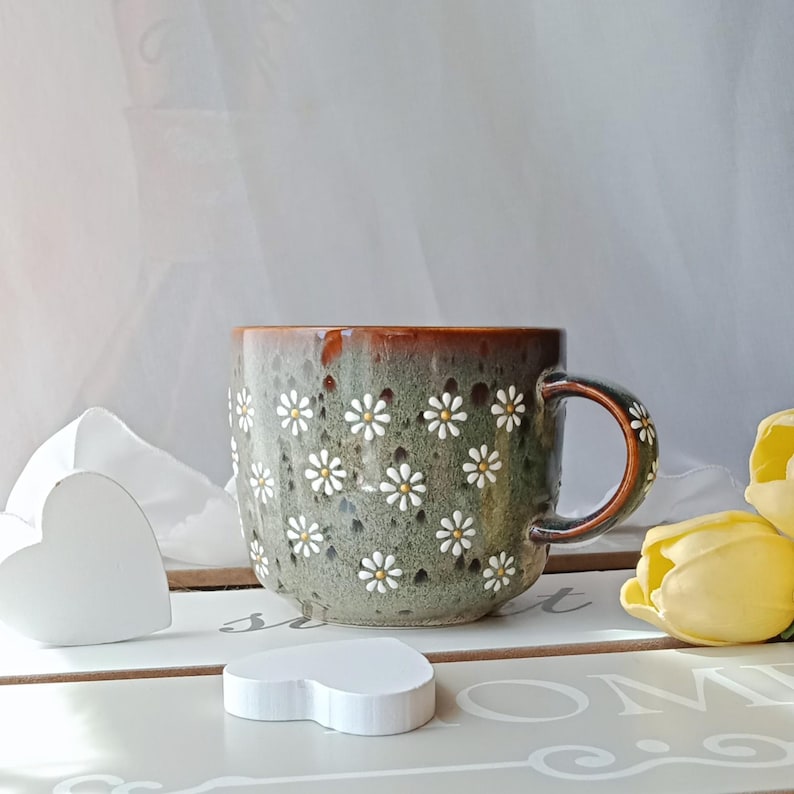 May include: A ceramic mug with a green and brown glaze, adorned with white daisy flower patterns. The mug has a brown handle and rim. The mug is sitting on a white surface with a white heart-shaped decoration and yellow tulips.