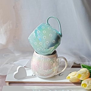 May include: Two ceramic mugs with a daisy pattern. The top mug is light blue with white daisies. The bottom mug is iridescent with pink and white daisies. The mugs are stacked on a wooden surface with a white heart and a sign that says "HOME".