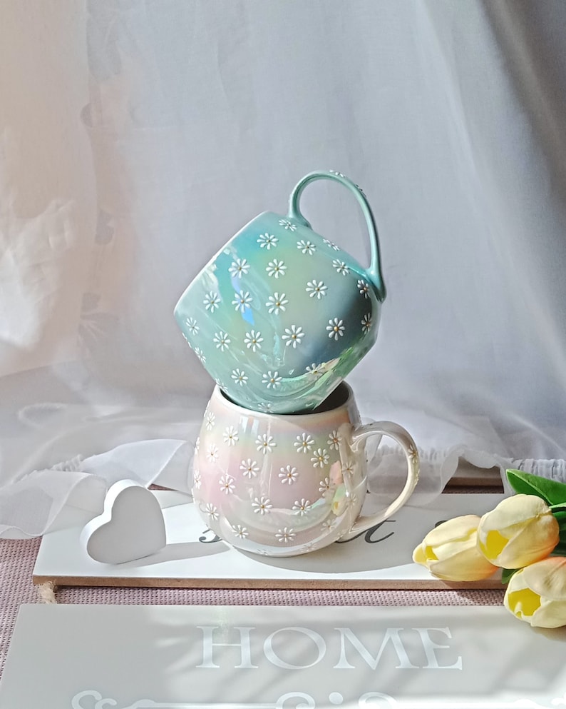 May include: Two ceramic mugs with a daisy pattern. The top mug is light blue with white daisies. The bottom mug is iridescent with pink and white daisies. The mugs are stacked on a wooden surface with a white heart and a sign that says "HOME".