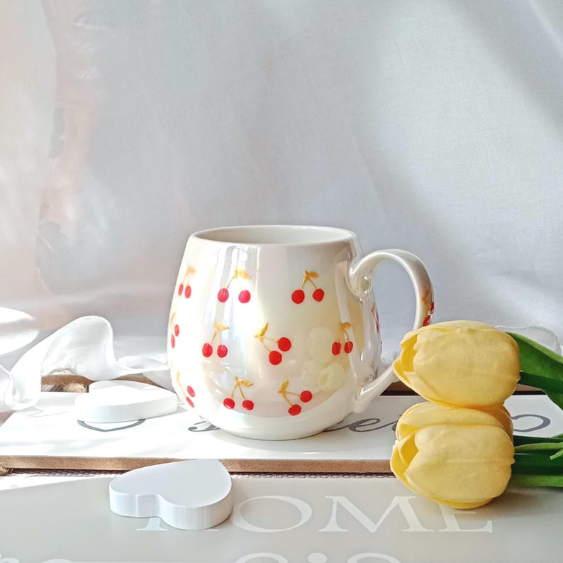 May include: Cream-colored ceramic mug adorned with a pattern of red cherries with yellow stems. The mug has a rounded shape, a curved handle, and a glossy finish. Two yellow tulips and heart-shaped objects are in the foreground.
