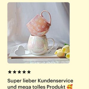 May include: Two pink ceramic mugs with a daisy pattern. The top mug is a lighter shade of pink and the bottom mug is a slightly darker shade of pink. The mugs are stacked on top of each other and are sitting on a wooden surface.
