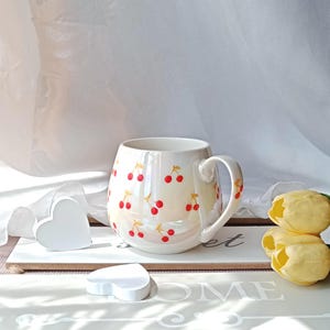 May include: White ceramic mug with a rounded shape and a handle, adorned with a pattern of red cherries. The mug is set on a white surface with heart-shaped decorations and yellow tulips.