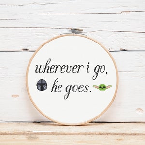 May include: White embroidery hoop with the text "wherever i go, he goes." The text is in black cursive font. A black and grey Mandalorian helmet and a green Baby Yoda character are embroidered on either side of the text.