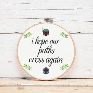 May include: A white embroidery hoop with a cross-stitch design featuring the text "i hope our paths cross again" and two green leafy sprigs on either side of the text. The text is in black cursive font. There are two small green and black pixelated images of a Star Wars character's helmet on either side of the text.