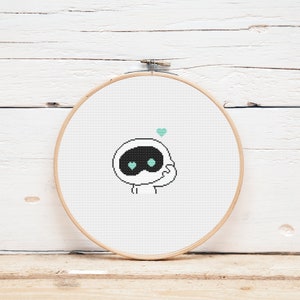 May include: A white cross-stitch embroidery hoop with a cute cartoon robot with black eyes and a teal heart above it.